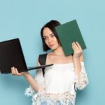 Widok z przodu młoda studentka w białej koszuli, niebieskie dżinsy i czarną torbę, trzymając laptopa i zeszyt na niebieskiej przestrzeni uniwersytet żeński