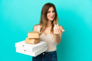 Kaukaski kobieta trzyma pizze i burger na białym tle na niebieskim tle zapraszając do przyjścia z ręką. Cieszę się, że przyszedłeś