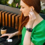Zbliżenie z góry uśmiechnięta młoda kobieta dotykająca bezprzewodowych słuchawek, słuchanie muzyki za pomocą telefonu komórkowego siedzącego przy stole, na tarasie kawiarni na świeżym powietrzu w letni dzień, rozmyte tło, selektywne focus