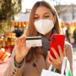 Stylowa kobieta w masce ochronnej KN95 FFP2 obserwująca numer karty kredytowej do wstawienia na smartfonie do płatności online z jarmarkami bożonarodzeniowymi w tle