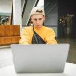 Skoncentrowany młody mężczyzna w jasnych, swobodnych ubraniach korzysta z laptopa w kawiarni w centrum handlowym z poważną miną
