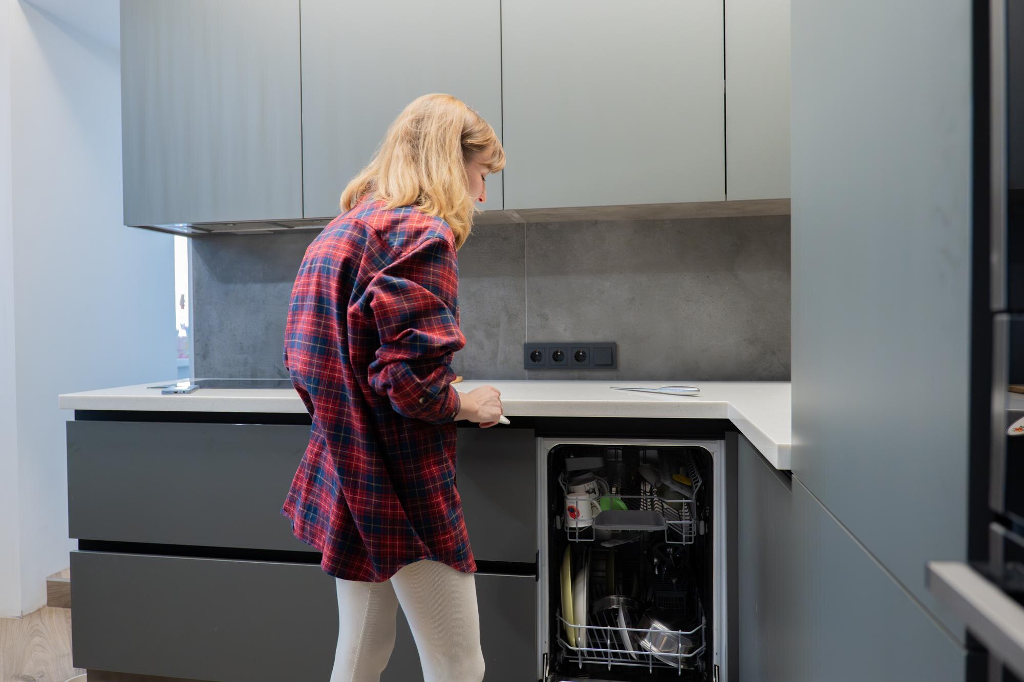 Young white woman loading dishwasher in contemporary gray kitchen wearing red plaid shirt focused on