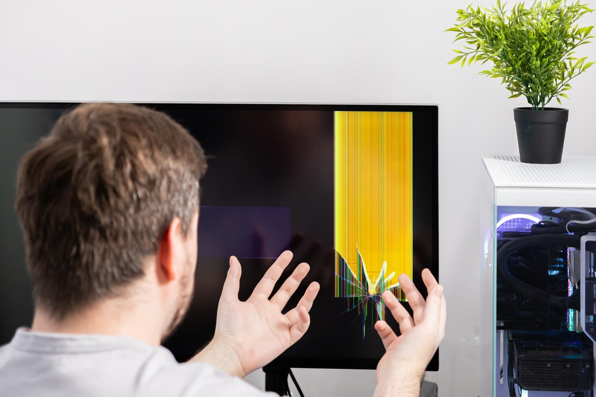 Frustrated person with hands reacting to a severely broken computer monitor displaying colorful lines signifying technical damage repair and unexpected expense