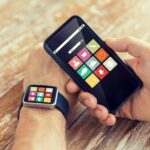 nowoczesna technologia, aplikacja i koncepcja ludzi - zbliżenie męskiej dłoni trzymającej smartfon i noszącej zegarek z ikonami aplikacji na ekranie w domu