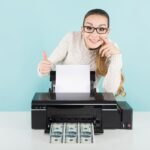 atrakcyjna szczęśliwa kobieta w swetrze i okularach gospodarstwa drukarki drukowania banknotów dolarowych z kciukiem do góry