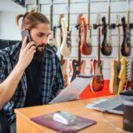 Poważny młody człowiek pracuje przy stole w pokoju. Patrzy na dokument, który ma pod ręką i rozmawia przez telefon. Wiele gitar elektrycznych wisi za nim.
