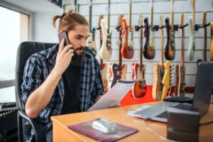 Poważny młody człowiek pracuje przy stole w pokoju. Patrzy na dokument, który ma pod ręką i rozmawia przez telefon. Wiele gitar elektrycznych wisi za nim.