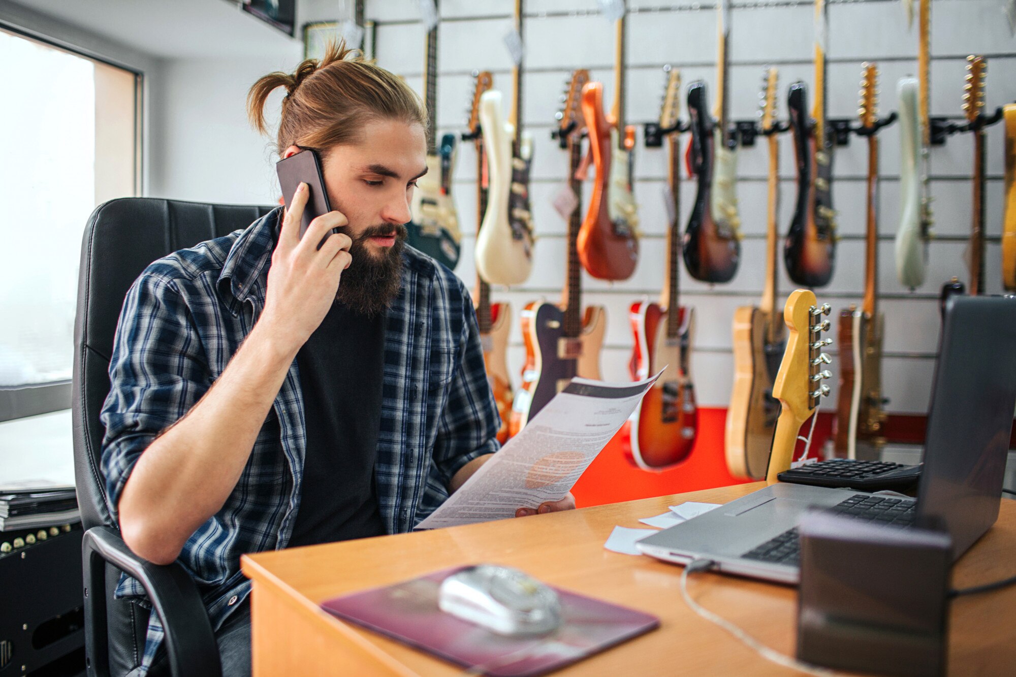Poważny młody człowiek pracuje przy stole w pokoju. Patrzy na dokument, który ma pod ręką i rozmawia przez telefon. Wiele gitar elektrycznych wisi za nim.