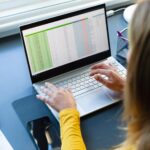 Biała kobieta siedząca przy biurku, pracująca z domu i używająca laptopa w słonecznym pokoju